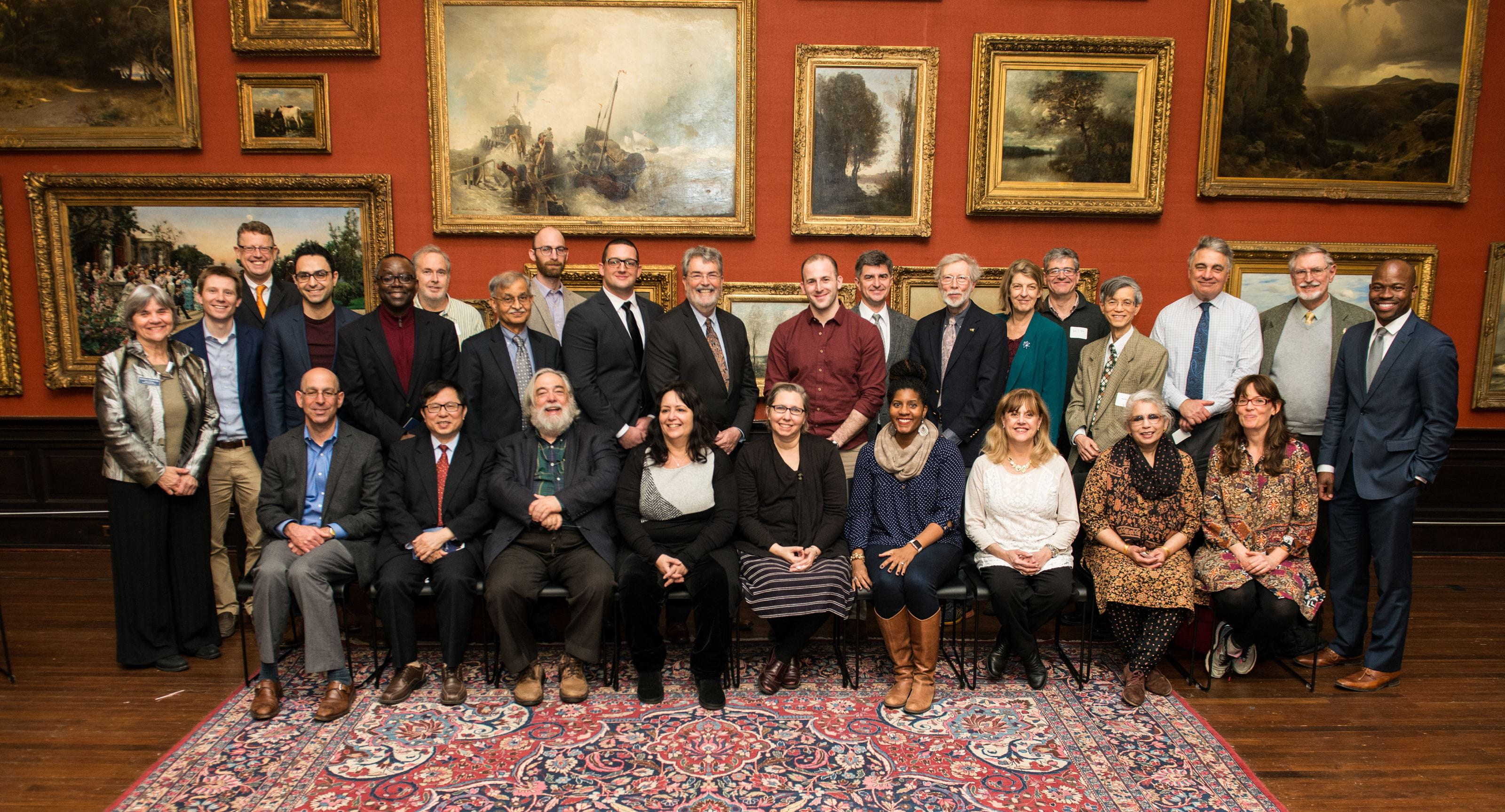 Men and women sit in two rows. They are posed in front of a wall with several large framed paintings. 