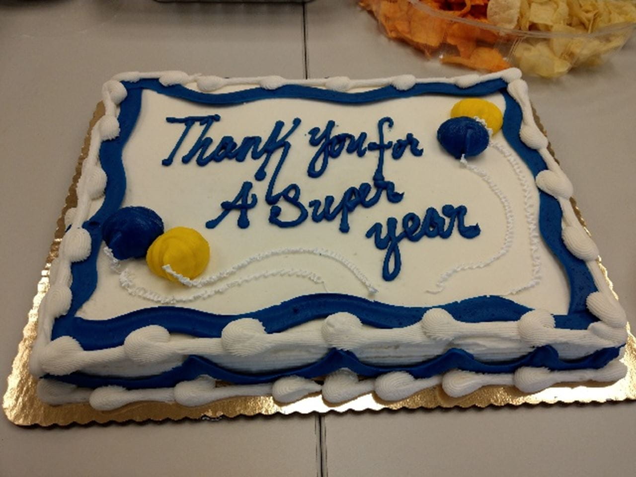 A cake with white icing and blue trip with yellow ballons. The cake says "Thank you for a super year"