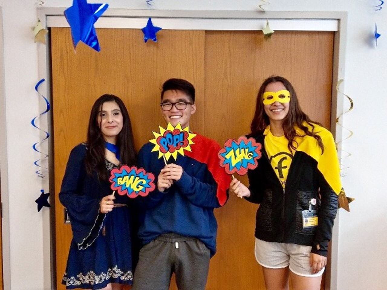 Three students stand next to each other. They hold colorful signs that say "pop!" "boom!" and "zap!"