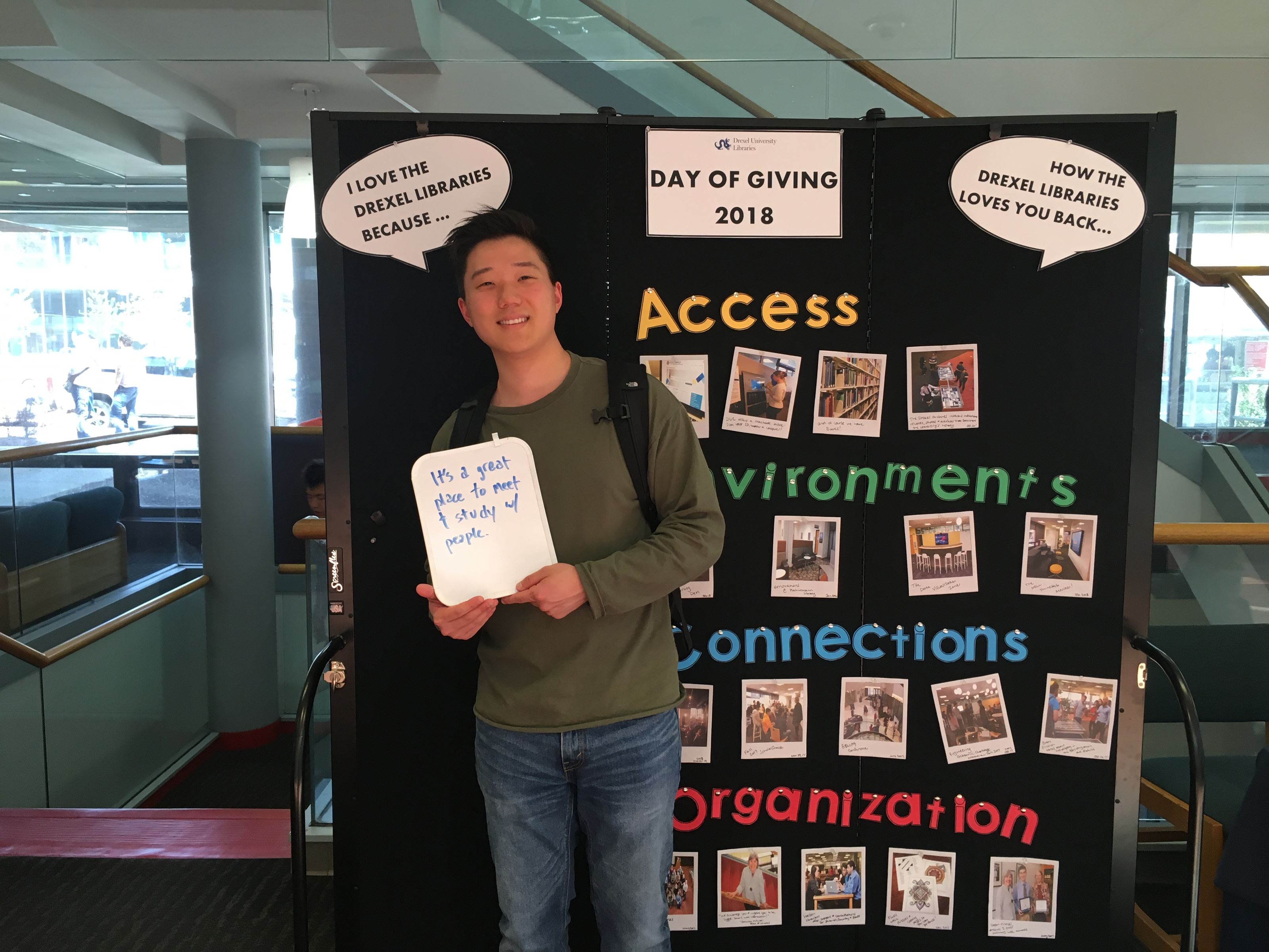 A young man holds up a dry erase board with writing on it. He is standing in front of a large board with photos on it. 