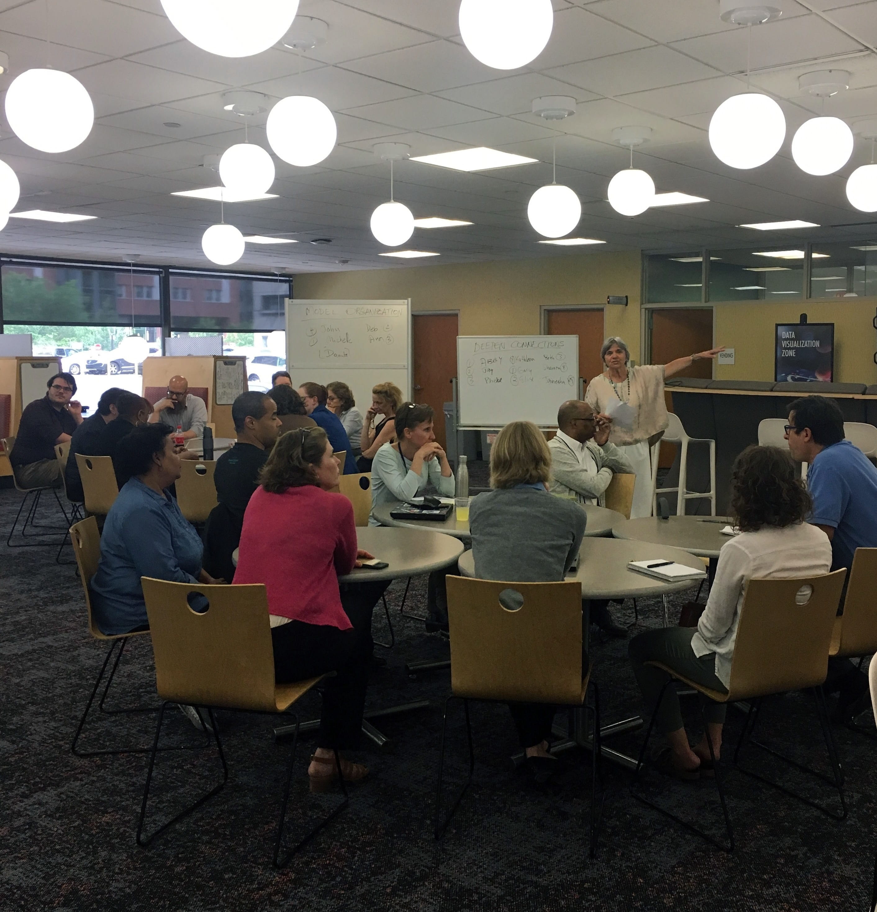 A group of people sit at round tables in a large while one woman speaks to them. 